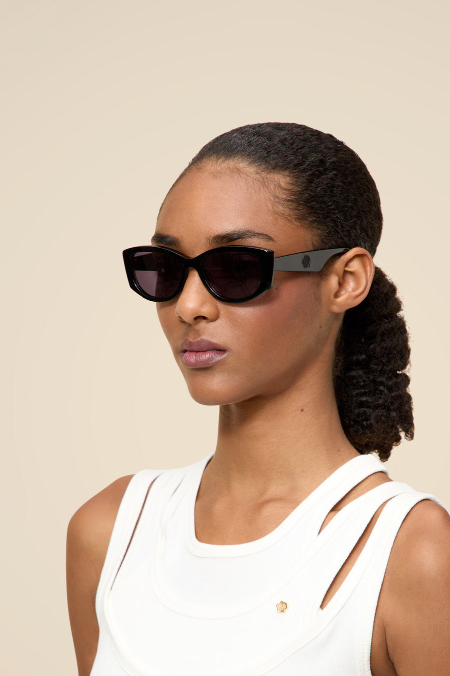 Sleek modern black rectangular sunglasses on woman with dark curly hair, wearing a stylish white top, against a neutral background.