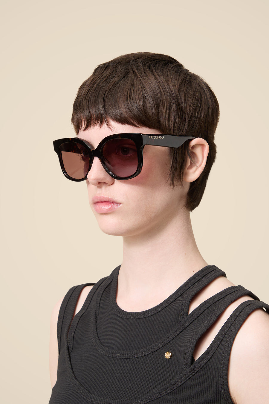 Short dark hair woman wearing oversized black sunglasses and a sleeveless ribbed black top against a beige background.