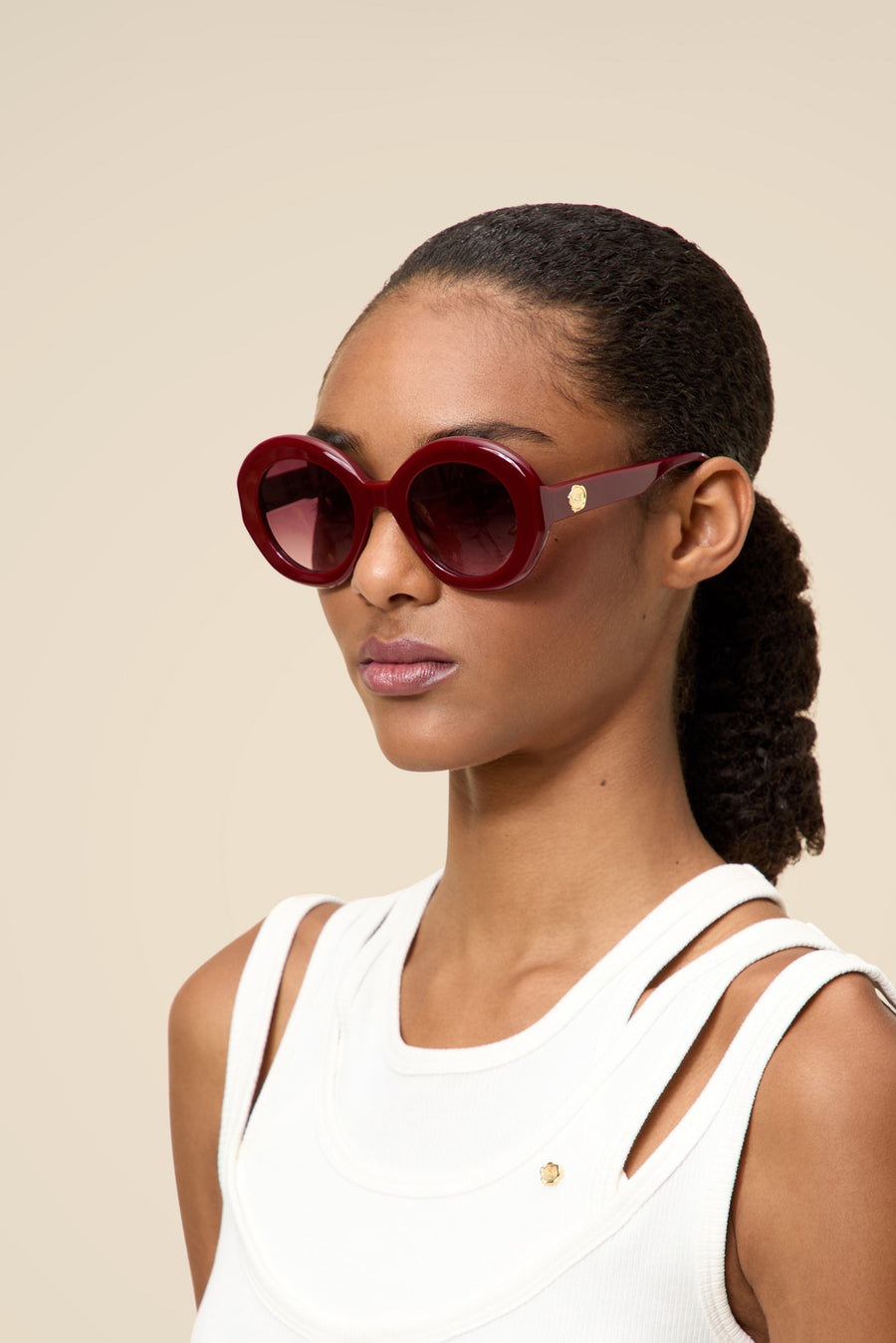 Round burgundy sunglasses with glossy frames and dark lenses, accented by a subtle gold detail, worn with a chic white top.