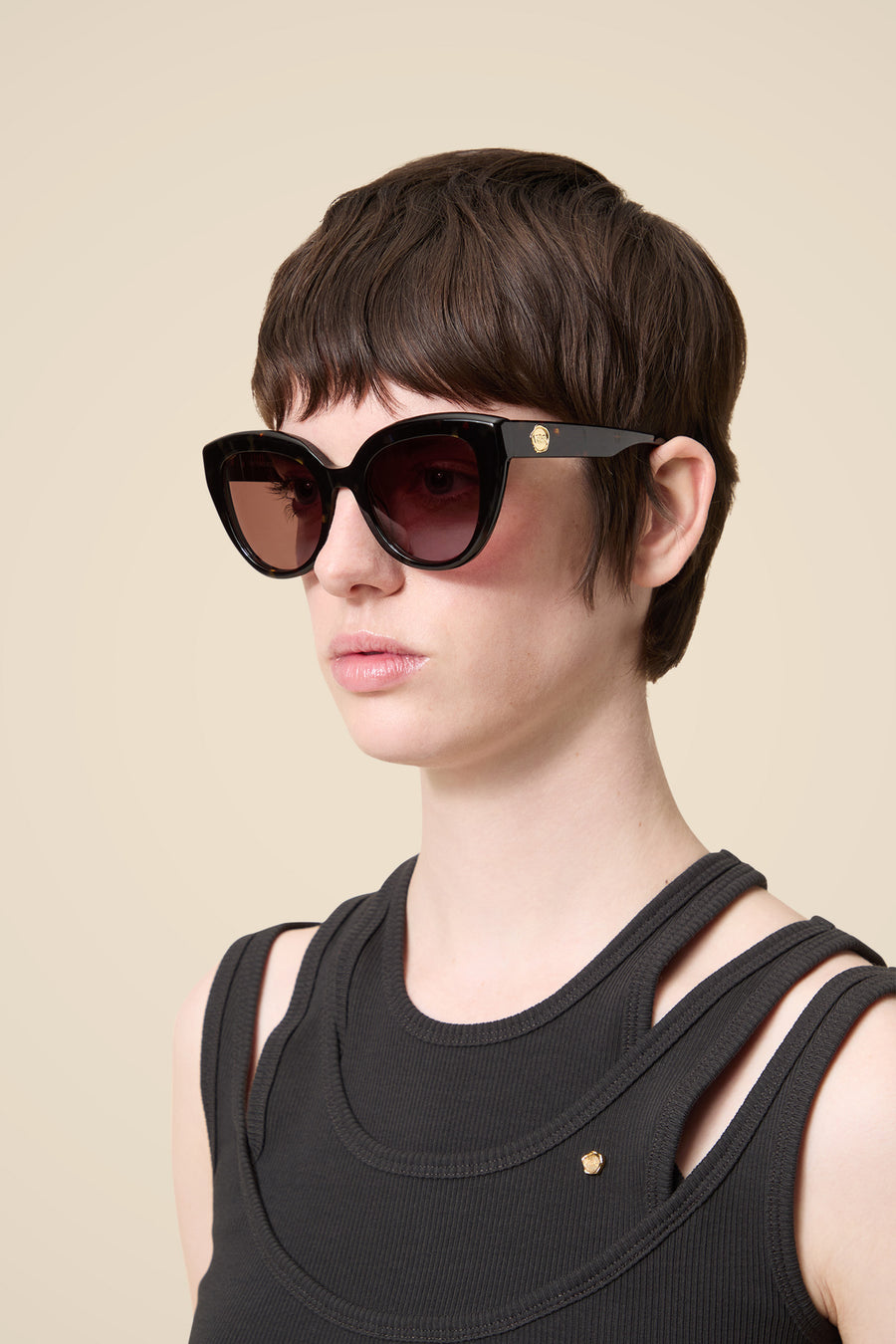 Black cat-eye sunglasses with dark gradient lenses, worn by a model in a sleeveless ribbed black top with gold detailing.