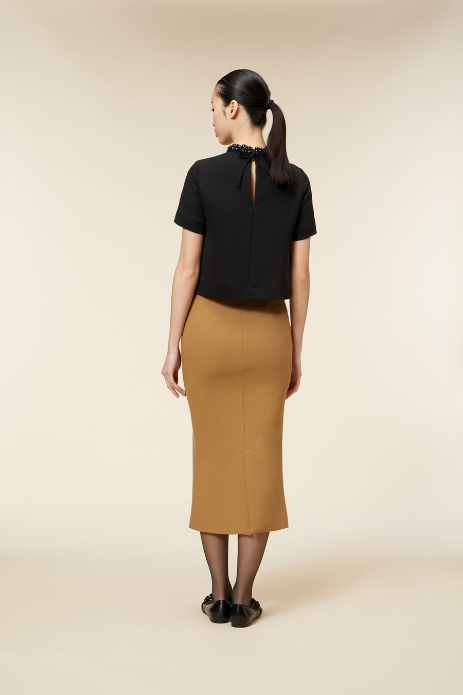 A chic black top features a keyhole back and bead detailing at the neckline, paired with a camel pencil skirt.