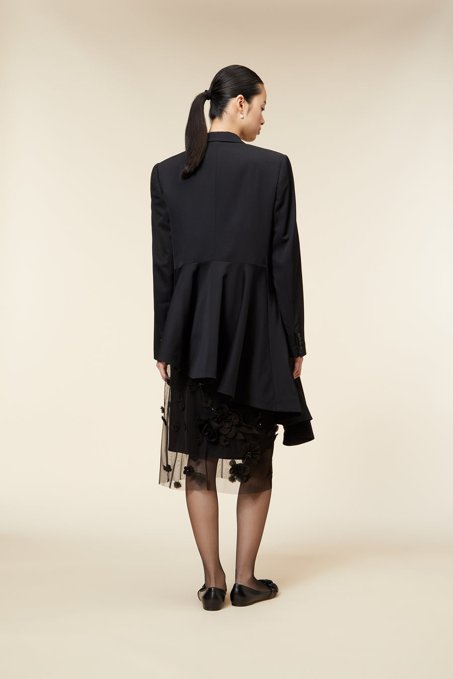 Model in a black asymmetric blazer over floral detailed mesh skirt, standing on a neutral background.