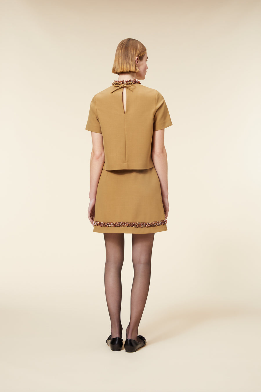 Model in a camel short-sleeve top with a cutout back, matching skirt with embellished hem, black flats, and sheer stockings.