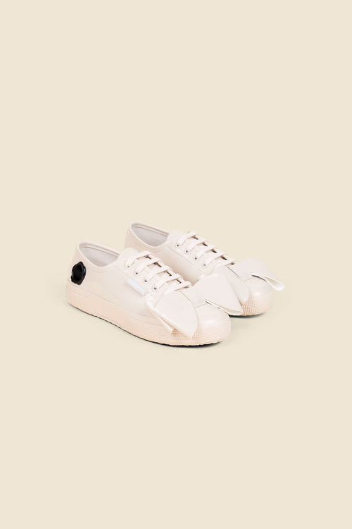 White patent leather sneakers with oversized bow detail and black rose embellishment, showcasing a minimalist design style.