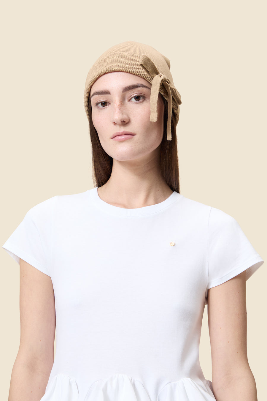 A stylish beige knit beanie with a tied bow detail worn with a simple white top, showcasing minimalist elegance.