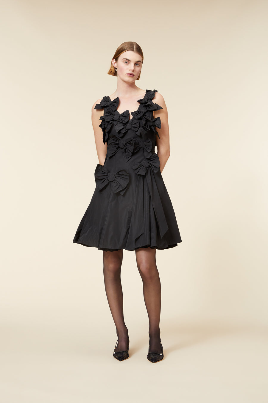 A black cocktail dress with oversized bow detailing, sleeveless design, and flared skirt, paired with sheer black stockings.