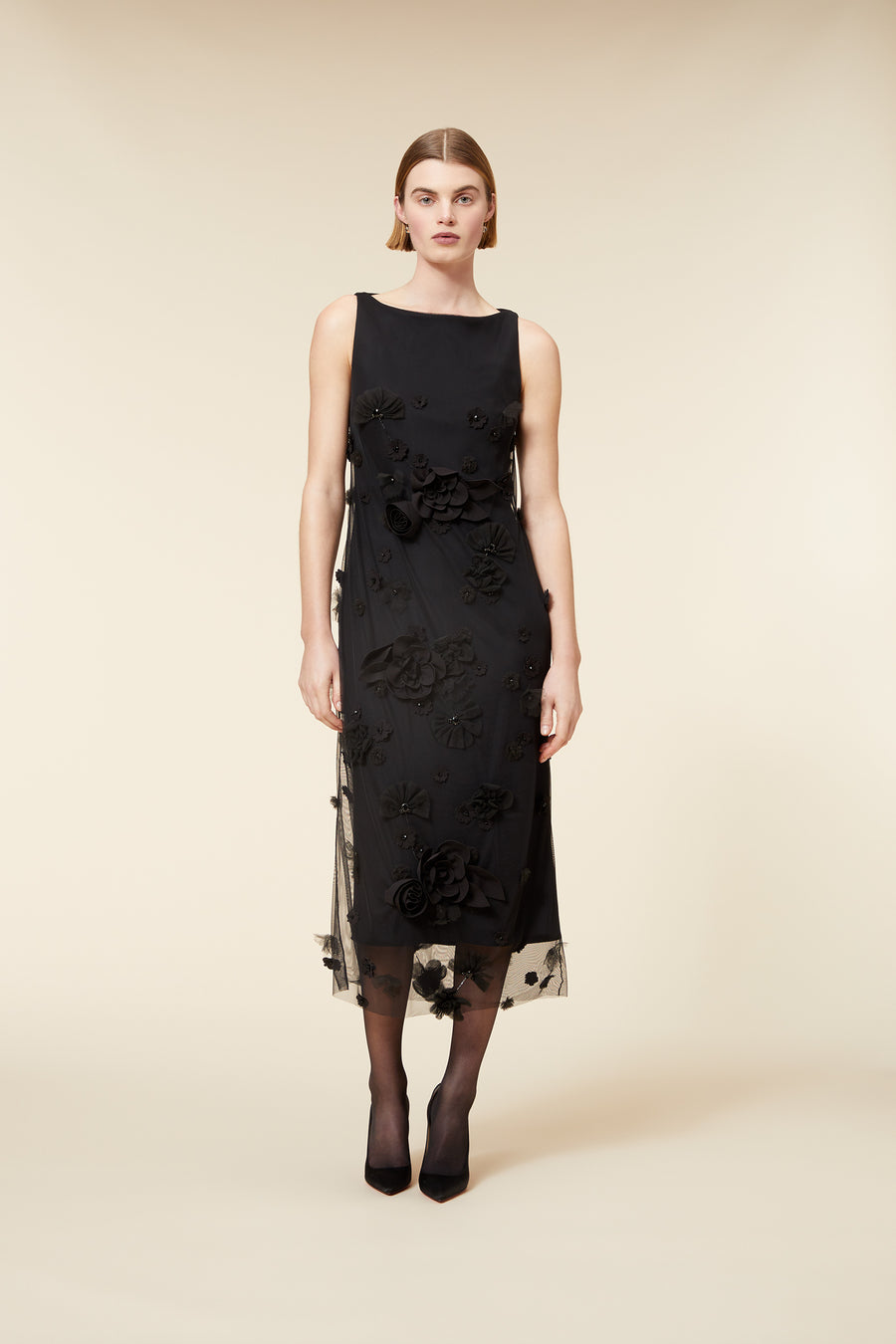 Elegant black sleeveless dress with intricate floral appliqué detailing, worn by a woman in a minimalist setting.