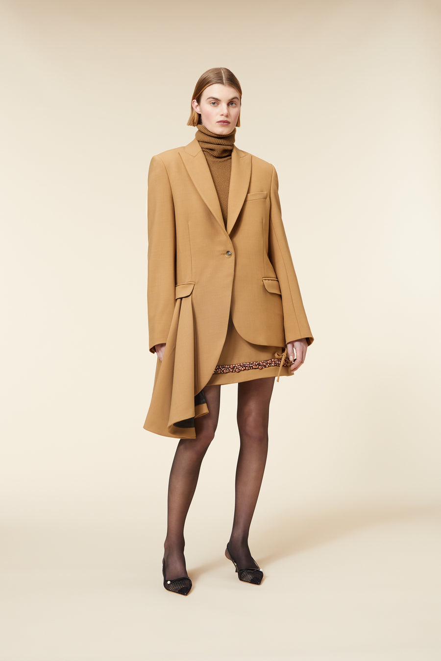 A model wearing a camel-colored asymmetrical blazer over a brown turtleneck, paired with a short skirt and black heels.