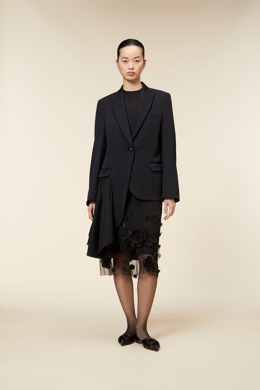 Model wearing a stylish black blazer over a knee-length black skirt with floral appliqués and sheer detailing.