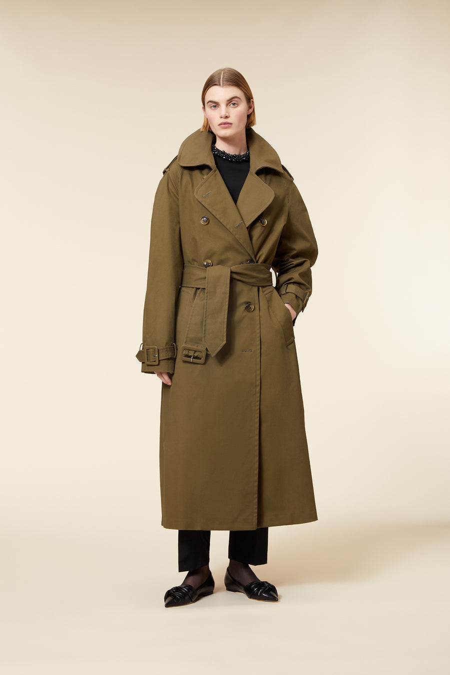 Olive green long trench coat featuring wide lapels, double-breasted buttons, and a belted waist, paired with black pants.