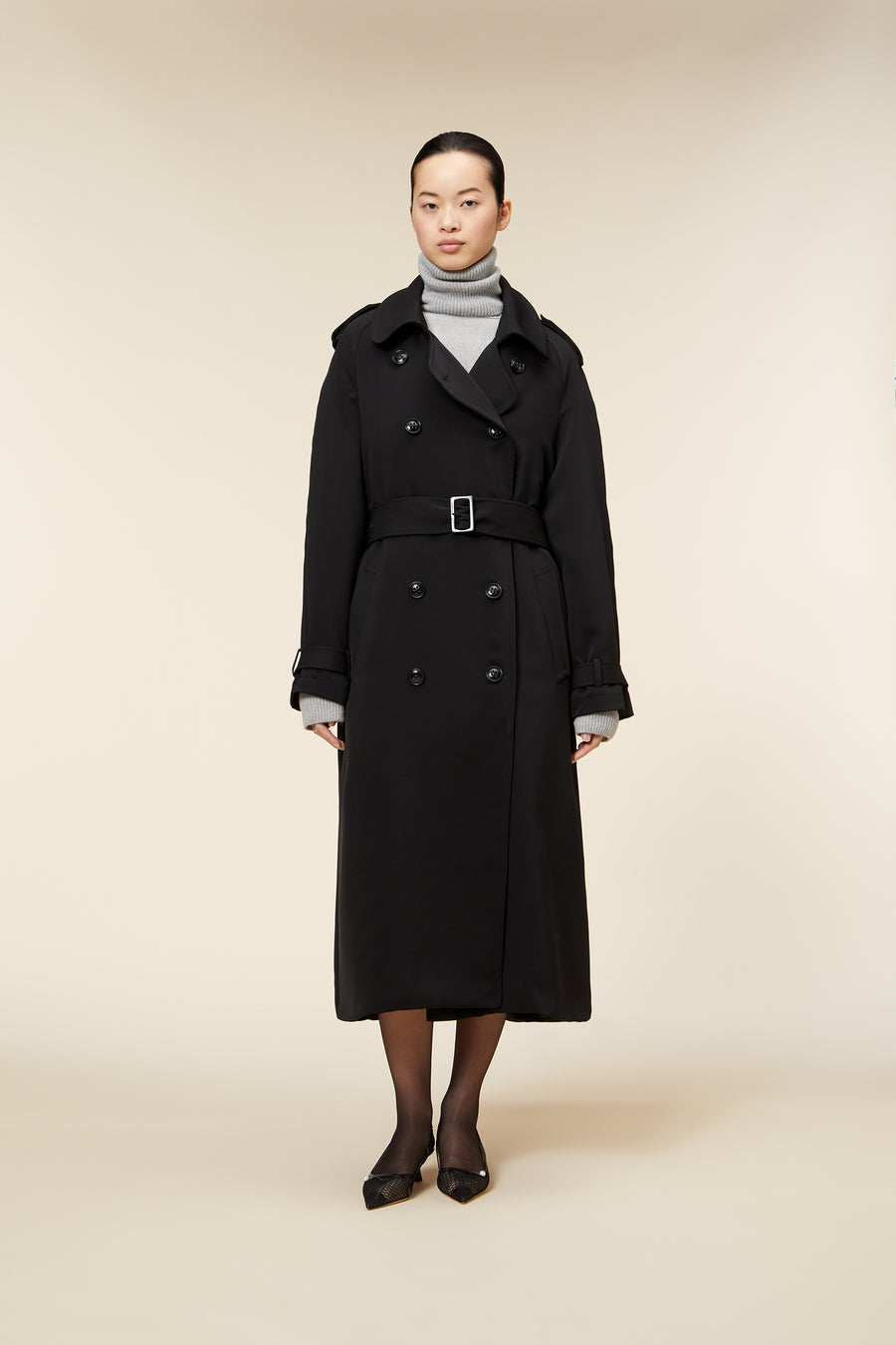 Long black trench coat with double-breasted buttons, belt, and notched lapel, worn over a gray turtleneck sweater.