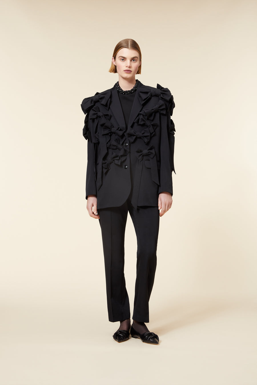 Elegant all-black suit with ruffled bow detailing, featuring tailored pants and paired with black loafers on beige background.