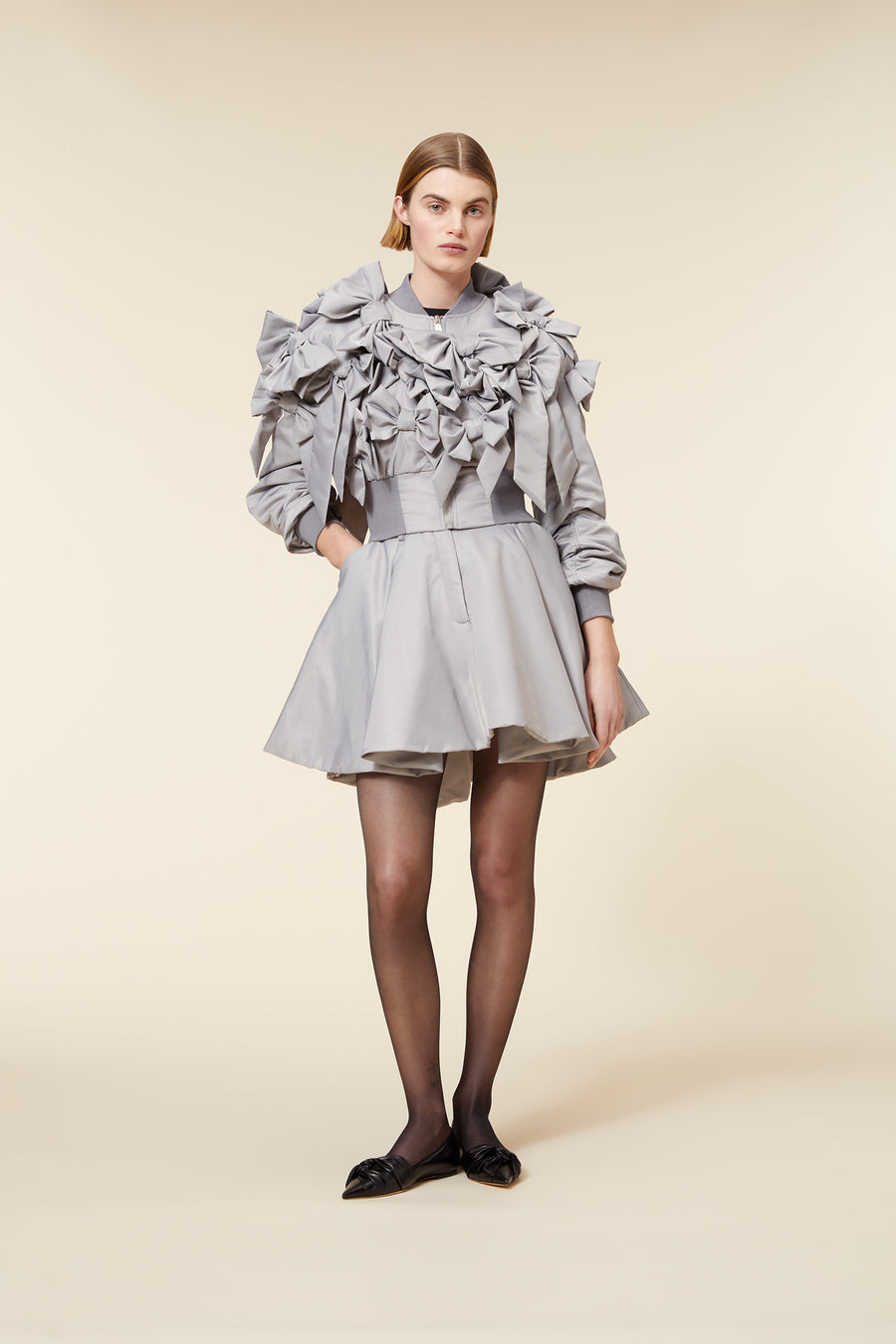 A model wears a gray structured dress with intricate ruffle detailing and puffed sleeves, styled with black pointed flats.