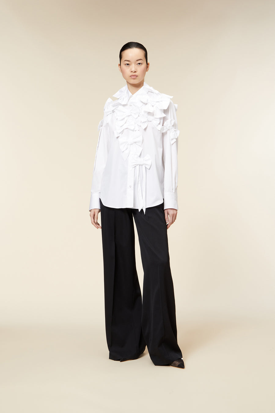 Model wearing a white ruffled blouse with bow details paired with black wide-leg trousers on a beige background.