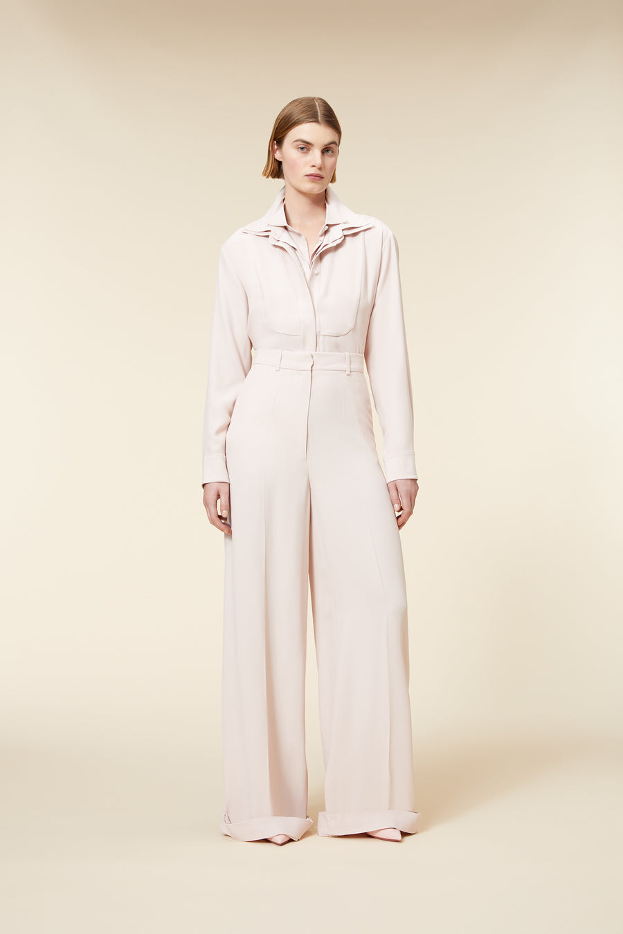 Woman wearing a light beige jumpsuit with wide-leg pants, collared neckline, and button-up front on a neutral background.