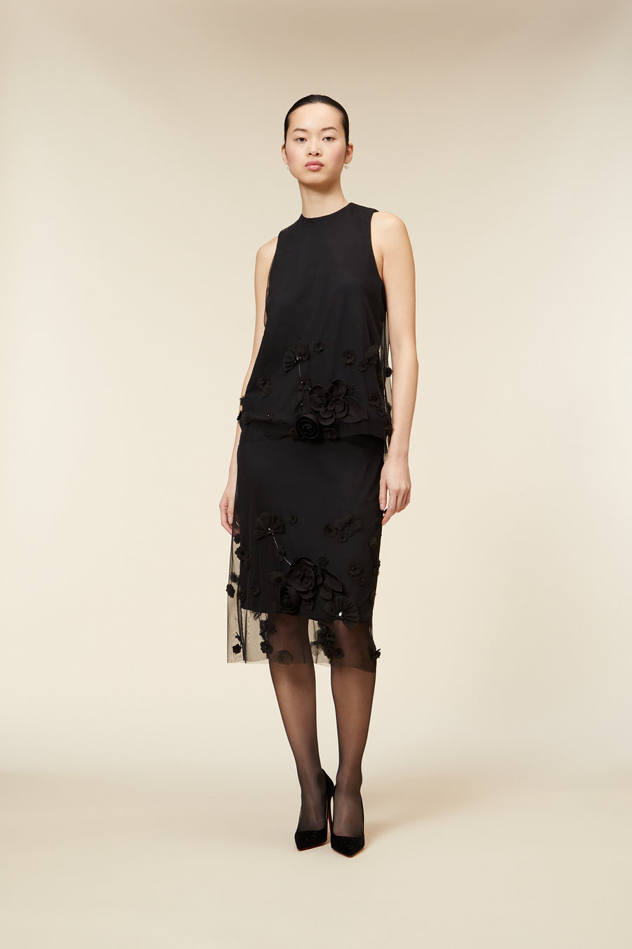 Elegant black dress with floral embroidery and sheer overlay on a woman standing against a neutral background.