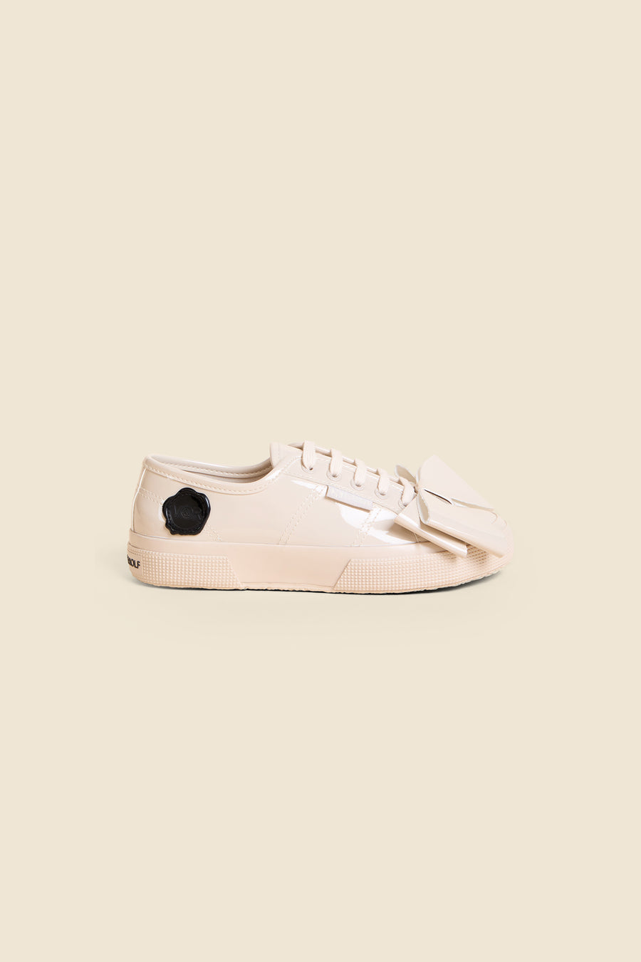 Beige glossy low-top sneaker with unique bow detail, textured sole, and black emblem on the heel against a neutral background.