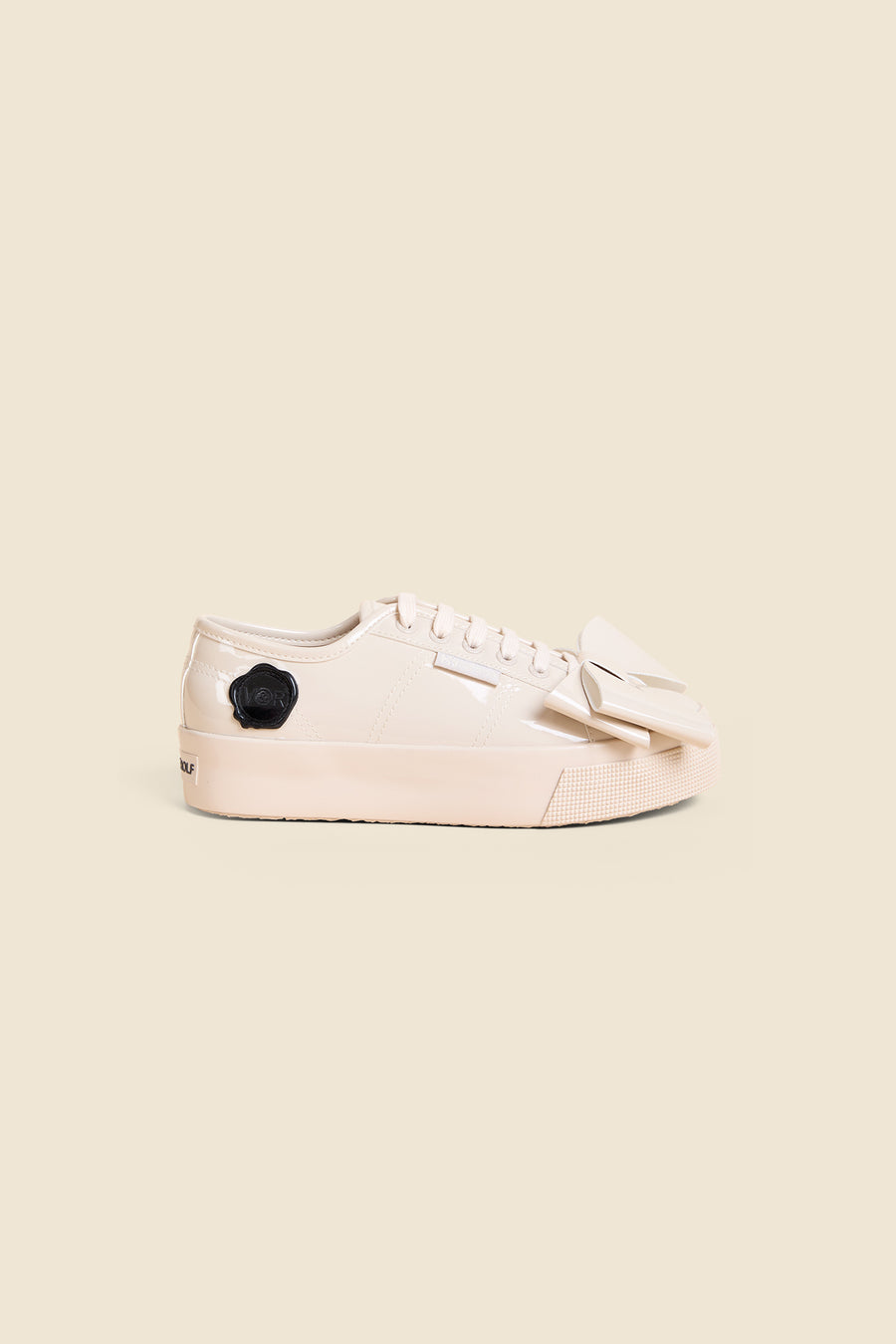 Cream patent leather sneaker with lace-up front and black logo detail, featuring chunky sole and unique angular bow accent.