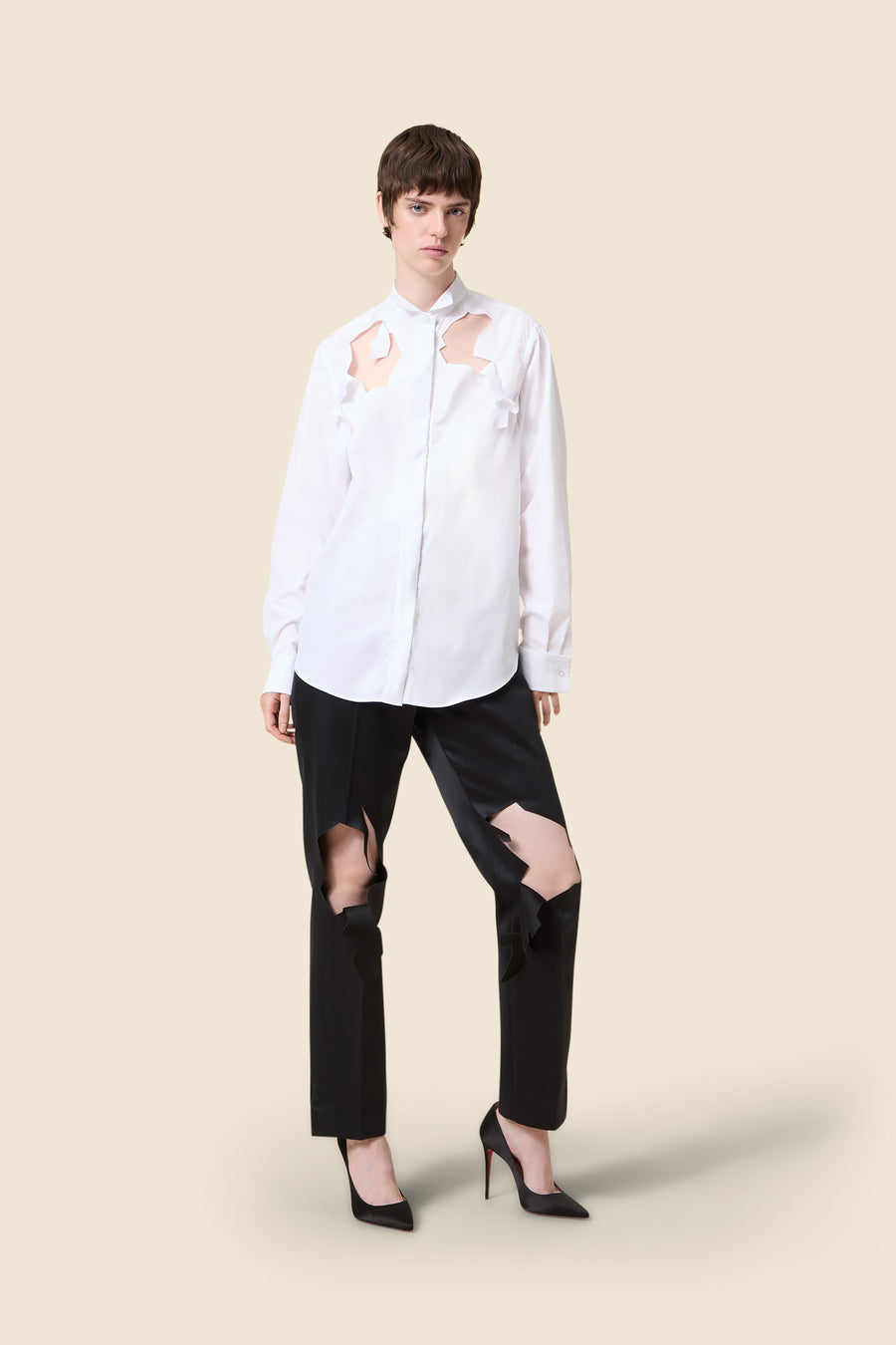 Fashion model in a white cutout blouse paired with bold black pants featuring unique cut designs, standing on a beige background.