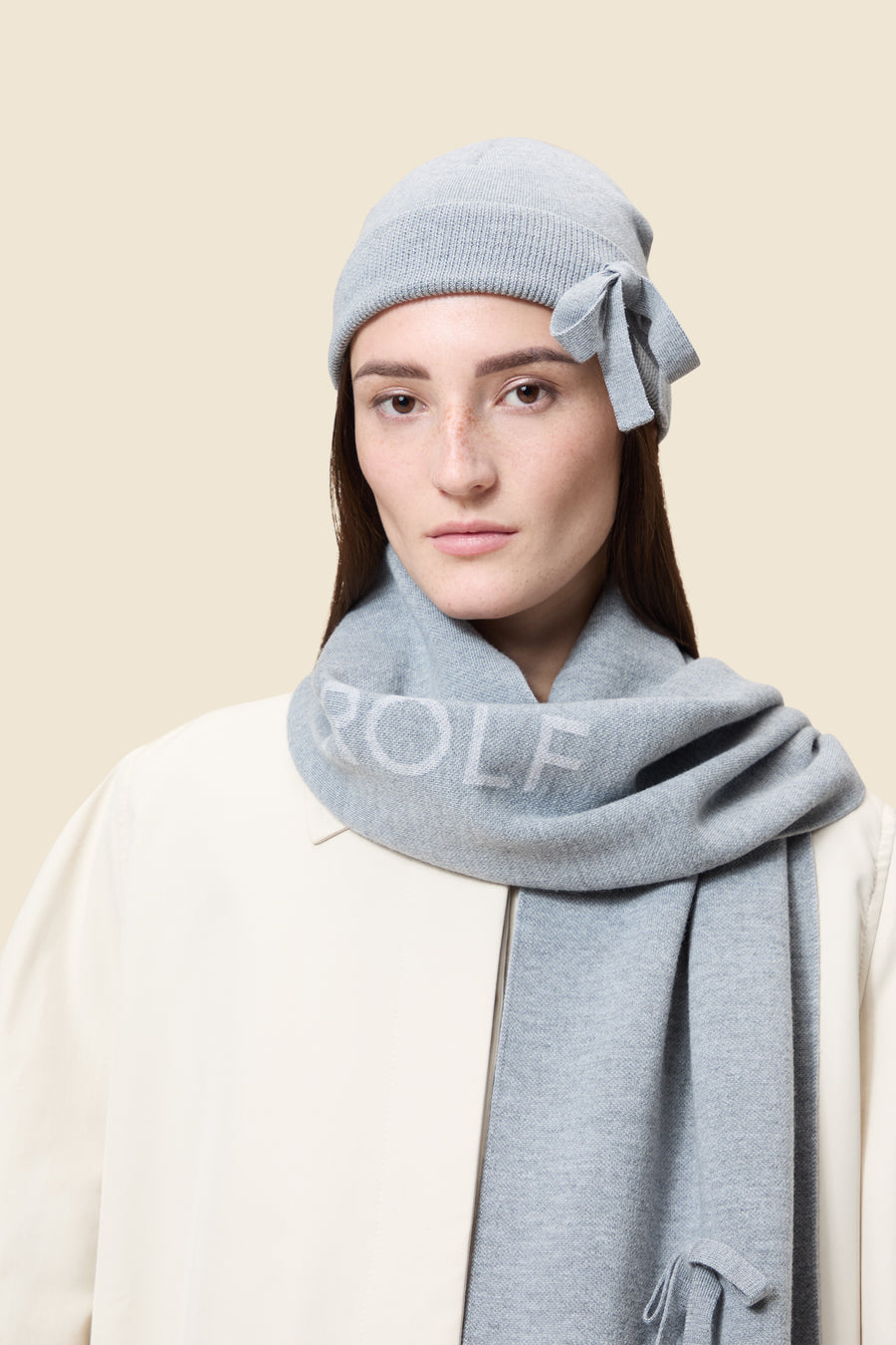 Light gray knit hat and scarf set with textured fabric and bow accent, worn over a cream coat background.