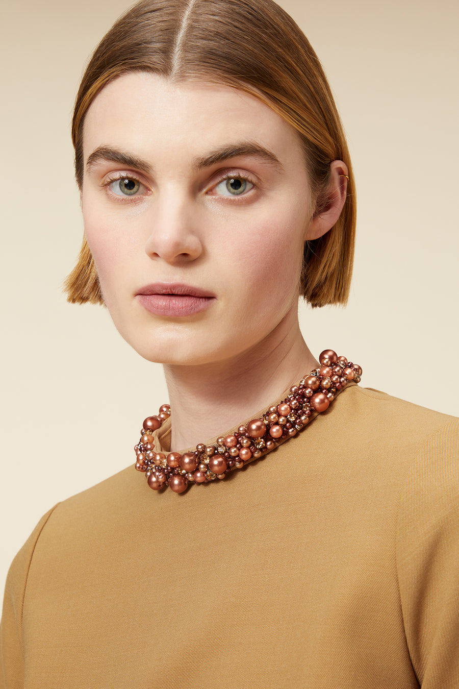 Woman wearing a beaded pearl necklace with copper tones, styled with a beige high-neck top in a minimalist setting.