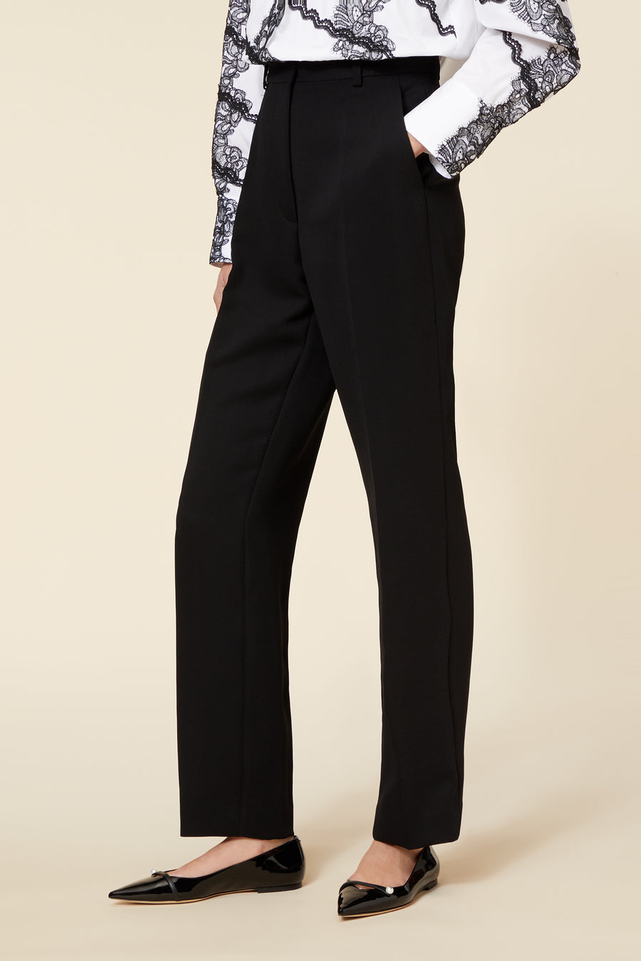 Black tailored trousers paired with a white lace-adorned blouse and glossy black ballet flats on a neutral background.