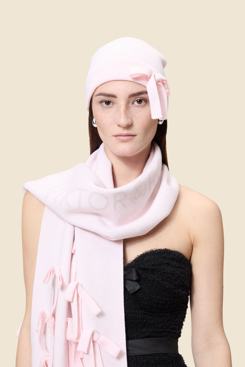 Pastel pink knit hat with bow detail paired with matching scarf, contrasting elegantly with a textured black dress.